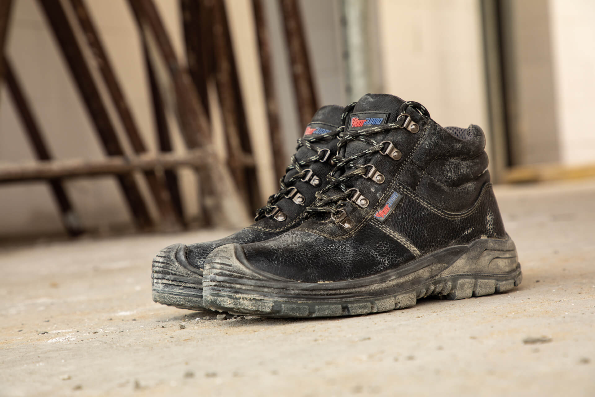 Auf dem Bild ist der stark beanspruchte, schwarze Sicherheitsschuh "Solid" von Footguard zu sehen. Die Schuhe stehen auf einem staubigen Betonboden in einer Baustellenumgebung und zeigen deutliche Gebrauchsspuren.