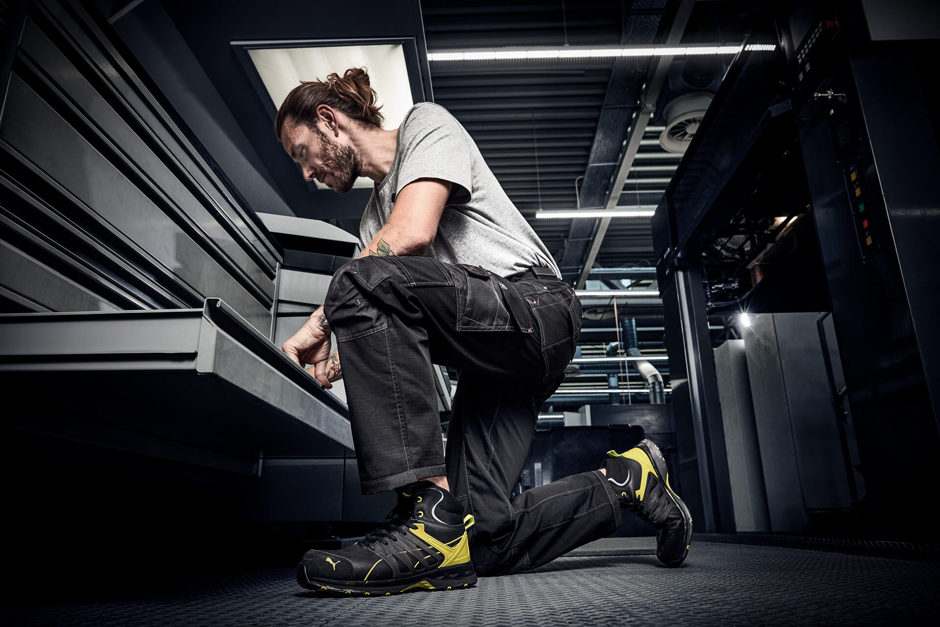 Ein Mann kniet in einer modernen Industrieumgebung vor geöffneten Schubladen eines Werkstattschranks. Er trägt schwarze Arbeitshose, graues T-Shirt und den schwarz-gelben Sicherheitsschuh "Velocity 2.0" von Puma Safety.