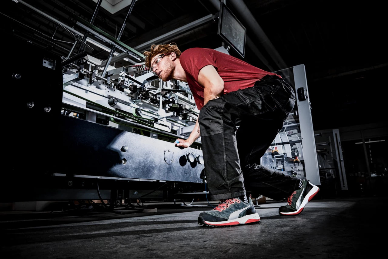 Ein Mitarbeiter bedient eine industrielle Maschine, während er den sportlichen Sicherheitsschuh "Speed" von Puma Safety in Schwarz-Rot-Weiß trägt.