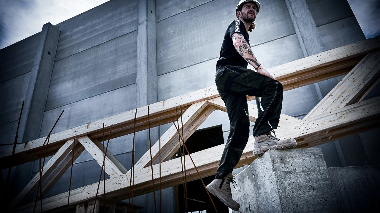 Ein Bauarbeiter trägt den Sicherheitsschuh "Conquest" von Puma Safety, während er auf einem Betonfundament vor einer Baustelle mit massiver Holzkonstruktion steht.