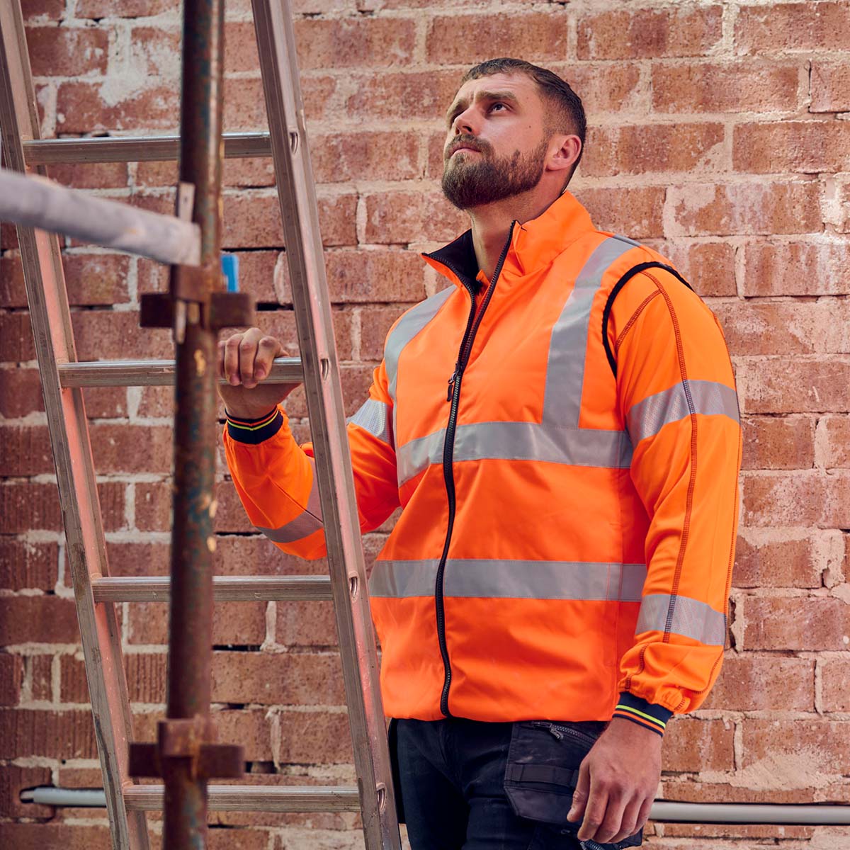 Handwerker trägt orange Bisley Warnschutzjacke mit Reflektorstreifen beim Arbeiten an einer Leiter – hochwertige Sicherheitskleidung für Baustellen.
