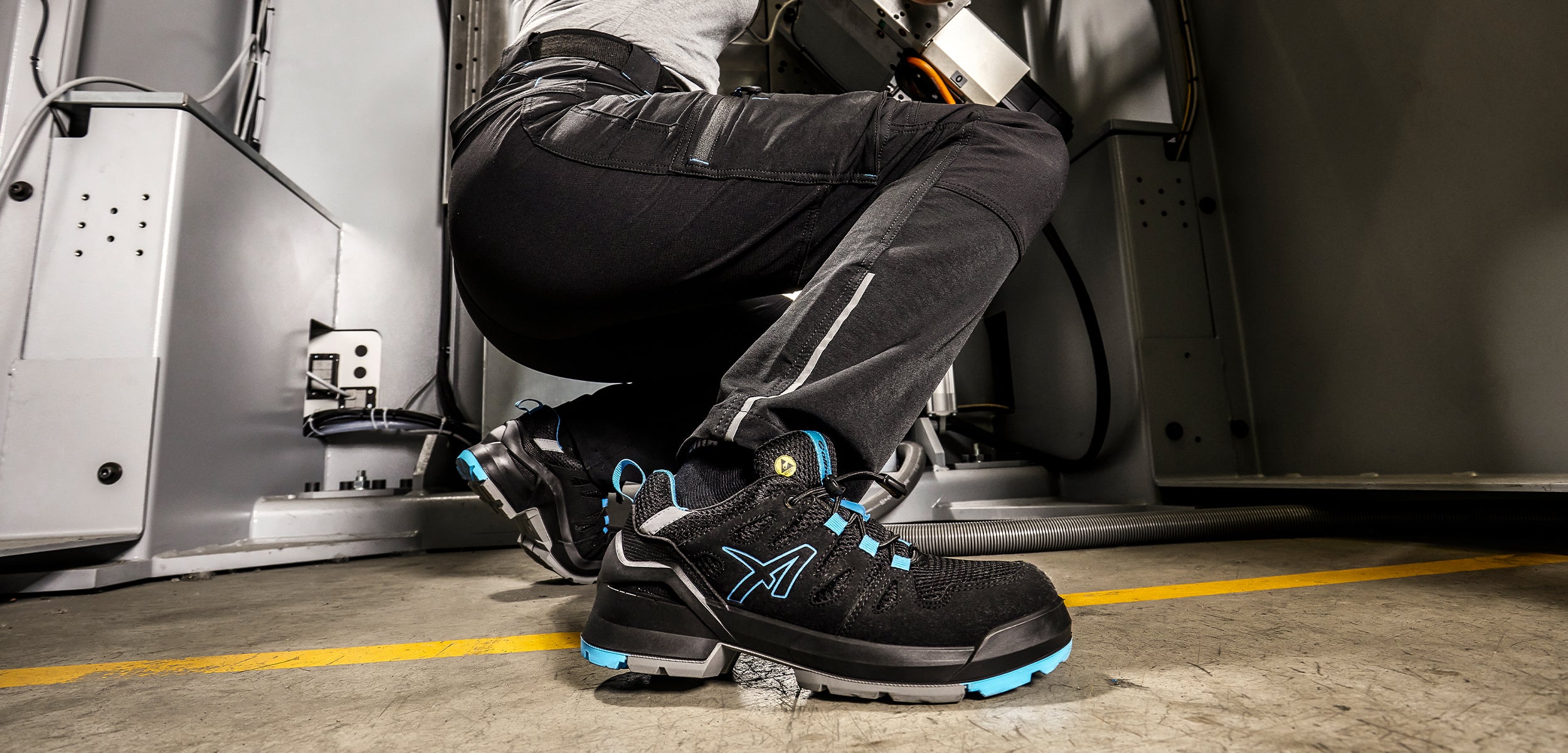 FASTPACK BLACK/BLUE LOW ALBATROS Sicherheitsschuhe S1PL ESD – schwarze Sicherheitsschuhe mit blauen Akzenten, getragen von einer Person bei der Arbeit in einer Werkshalle