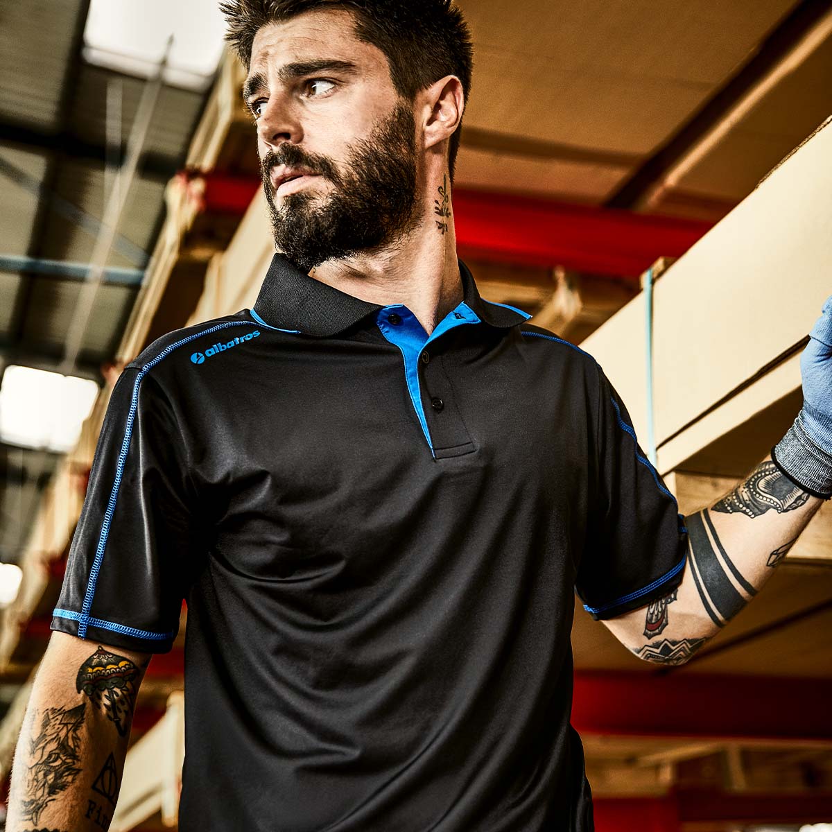 Mann im schwarzen Albatros Arbeits-Poloshirt mit blauen Akzenten beim Heben in Lagerumgebung - atmungsaktives T-Shirt für den professionellen Arbeitseinsatz im Innenbereich.