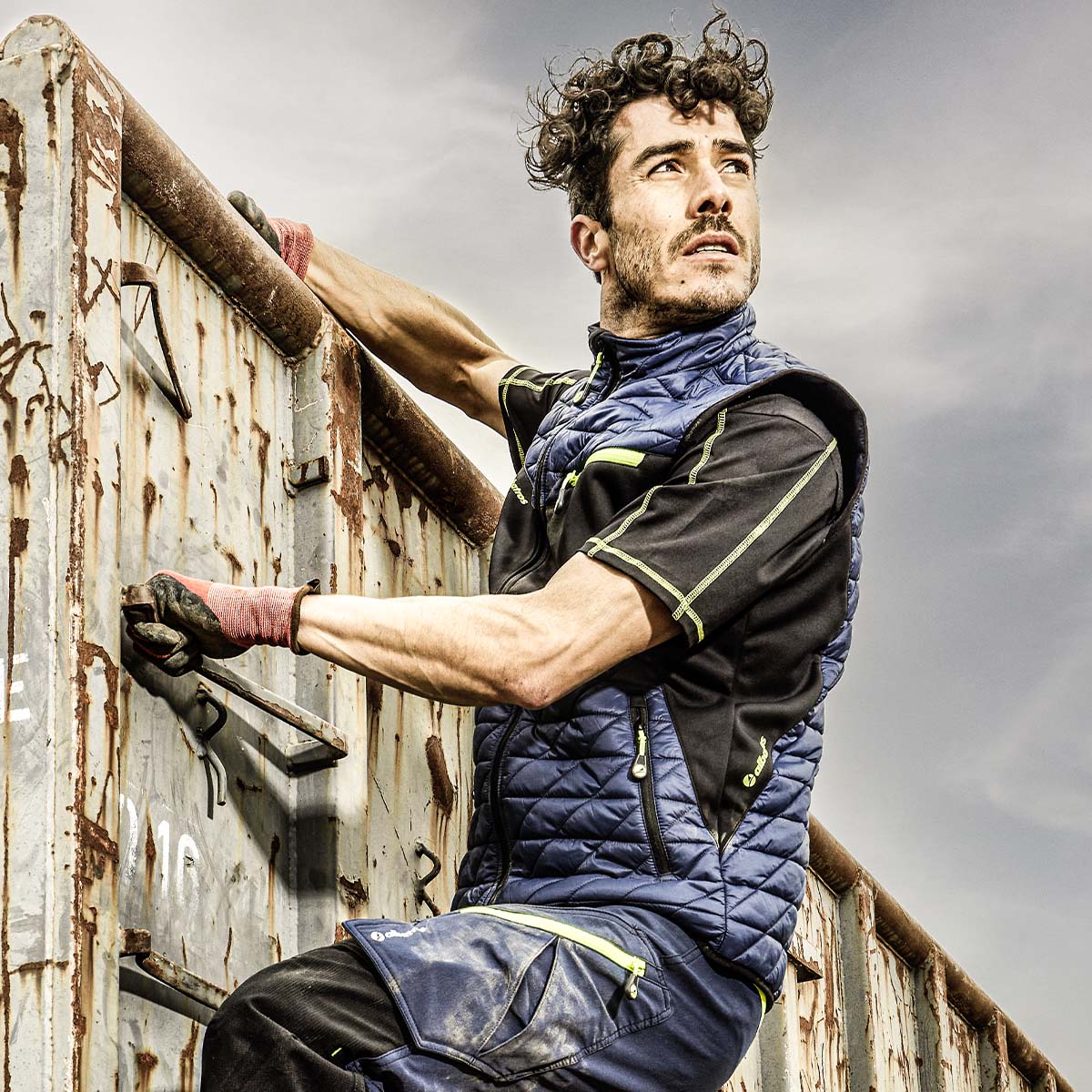 Arbeiter mit wattierter blauer Arbeitsweste und Funktionsshirt auf der Arbeit an einem rostigen Container - Outdoorkleidung für körperliche Einsätze im Außenbereich.