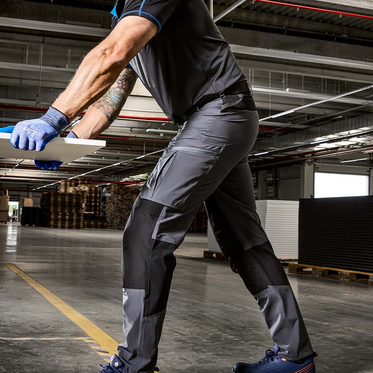 Graue Arbeitshose von Bisley mit Seitentaschen, getragen von einem Arbeiter beim Heben in einer Industriehalle - funktionale Berufsbekleidung im Einsatz.
