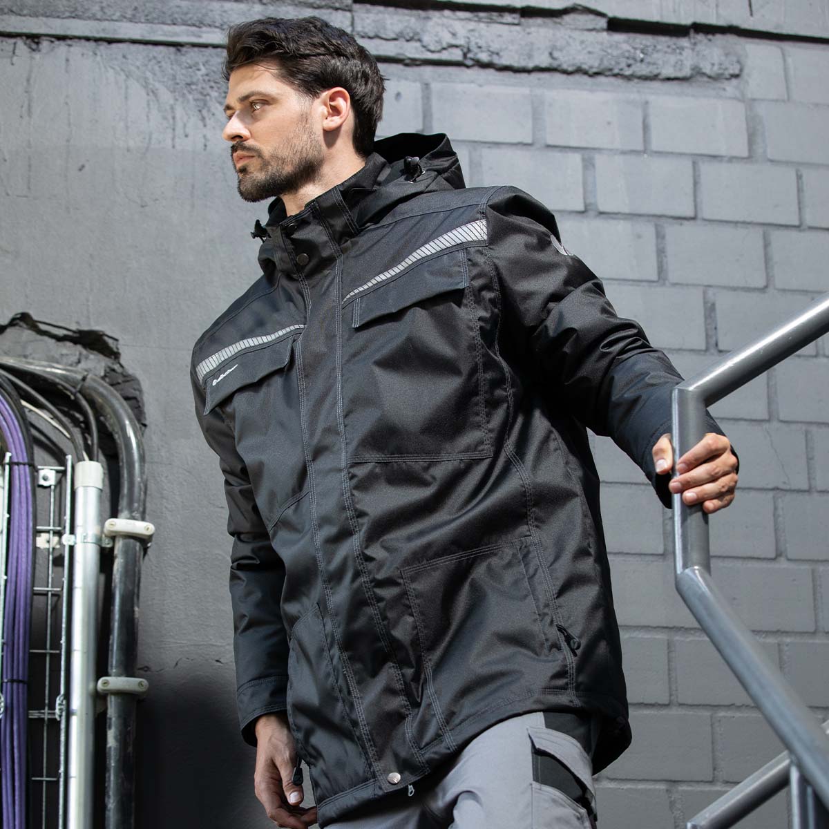 Mann in schwarzer Arbeitsjackemit reflektierenden Elementen und Kapuze auf einer Außentreppe vor einem Industriegebäude.