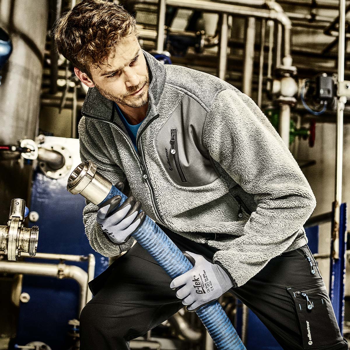 Graue ALBATROS® Fleecejacke mit Reißverschlusstaschen, getragen bei der Arbeit mit einem Schlauchsystem in industrieller Umgebung.