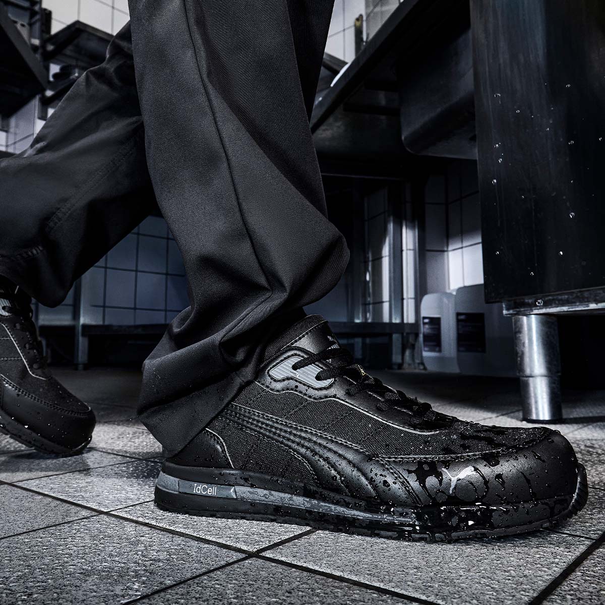 Schwarzer rutschfester PUMA SAFETY® Berufsschuh mit Wassertropfen auf der Oberfläche, getragen auf nassem Fliesenboden in einer Küche.