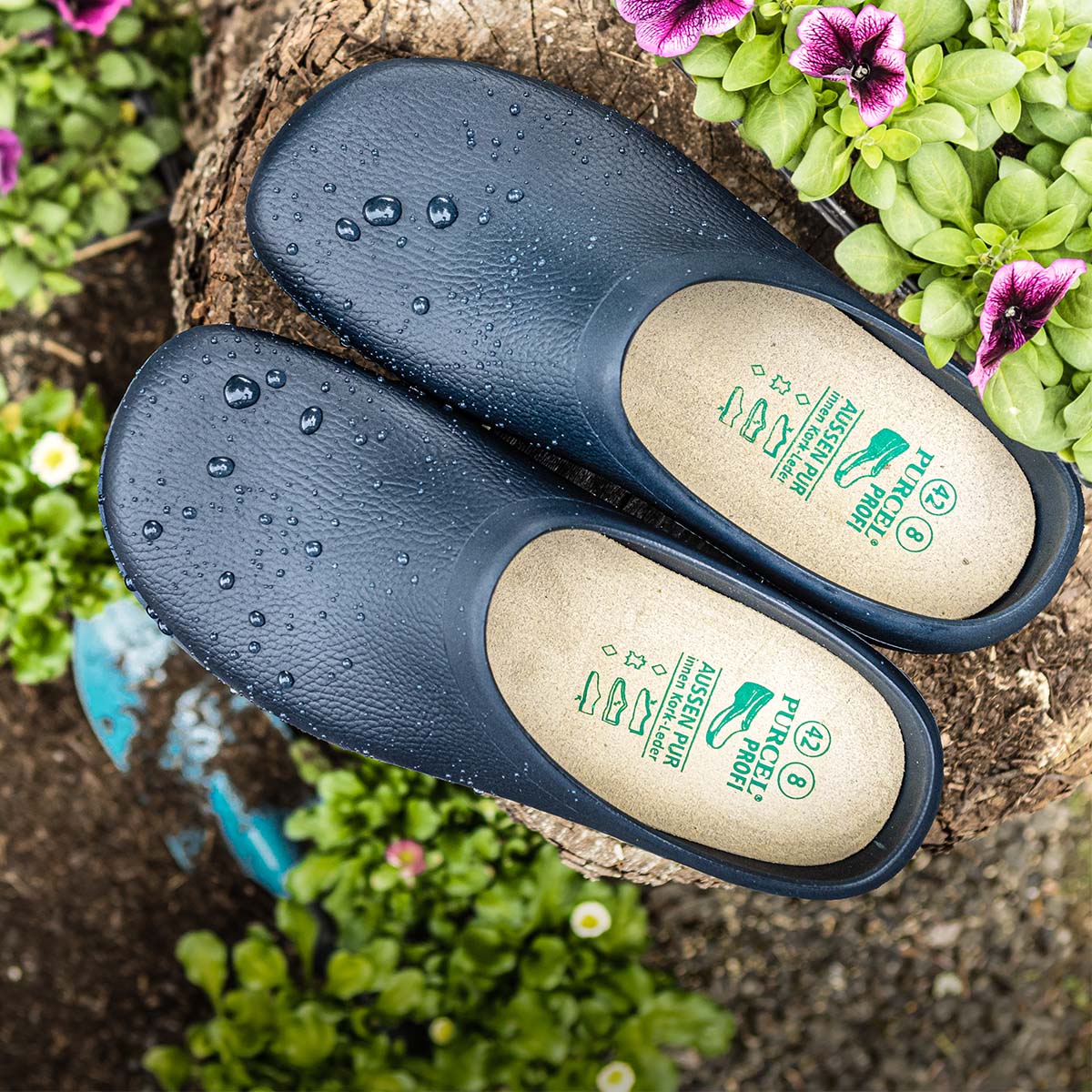 Dunkelblaue ALBATROS® Gartenclogs mit Wassertropfen auf der Oberfläche, fotografiert auf einem Baumstumpf zwischen blühenden Pflanzen.