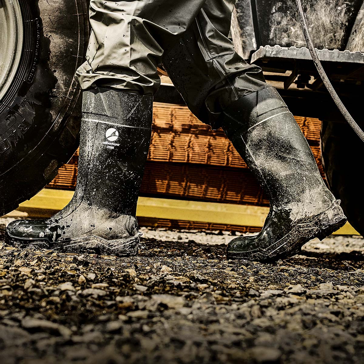 Verschmutzte ALBATROS® TITAN Gummistiefel, getragen auf nassem Schotterboden neben einem Traktorreifen.