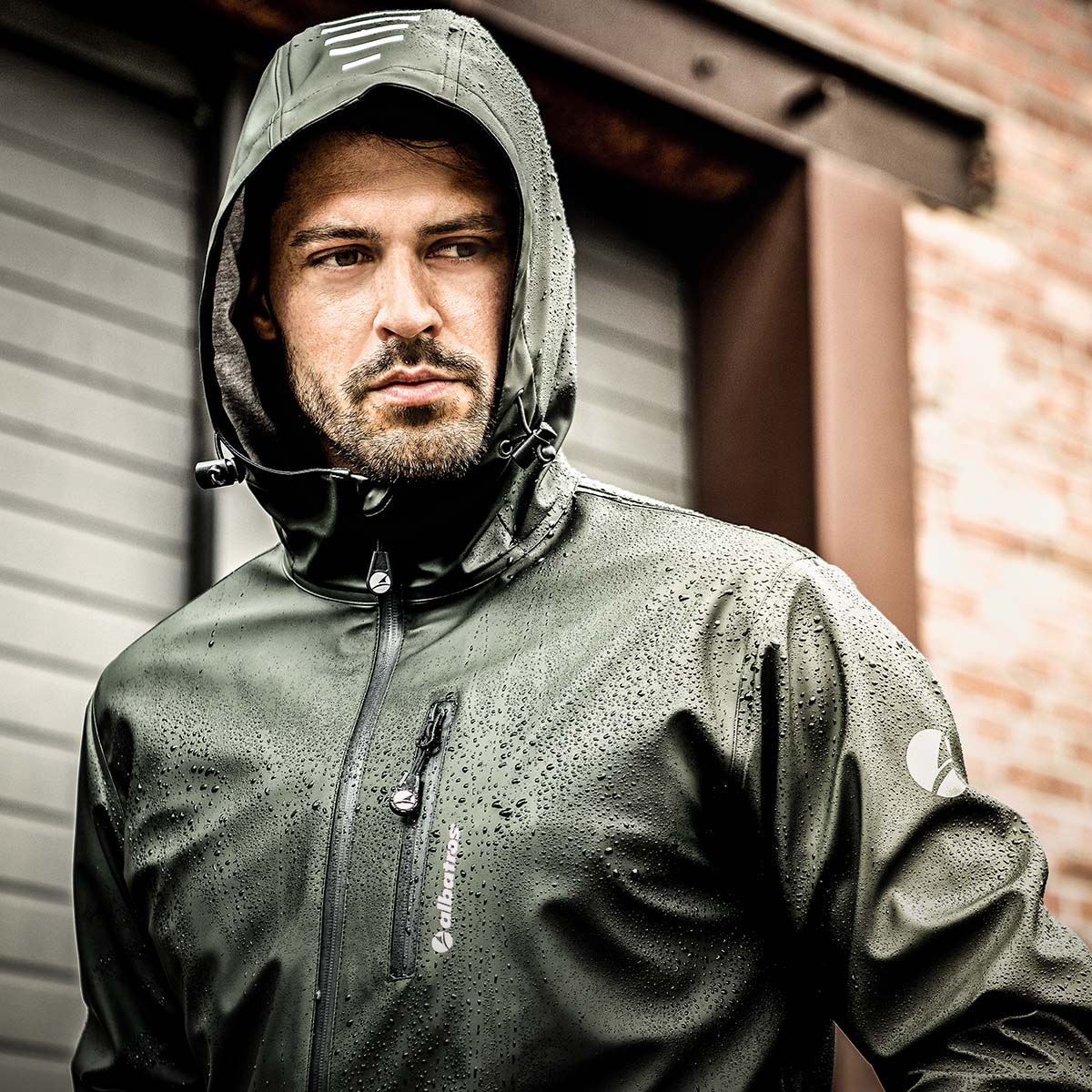 Olivefarbene ALBATROS® Regenjacke mit Kapuze und sichtbaren Regentropfen, getragen im Außenbereich vor einem Gebäude mit Rollladen.