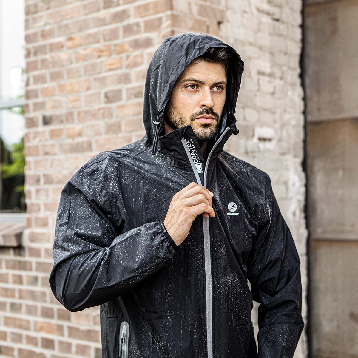 Wasserdichte schwarze Regenjacke von Albatros mit Kapuze und Reißverschluss, getragen von einem Mann bei Regenwetter - Outdoor-Arbeitskleidung mit reflektierendem Detail von COA.TEX vor einer Backsteinwand.