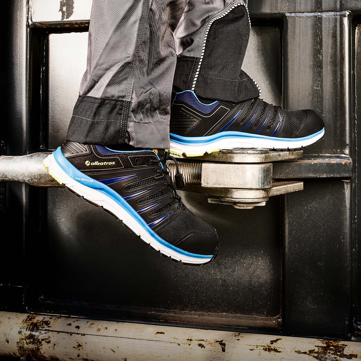 Schwarze ALBATROS® Schuhe mit blauen Akzenten und weißer Sohle, getragen auf einem Metallrohr vor einem dunklen Industriehintergrund.