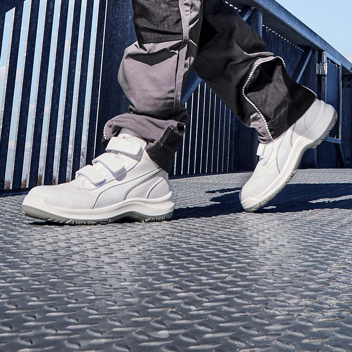 Weiße PUMA SAFETY® Schuhe mit drei Klettverschlüssen, getragen auf einem Gittersteg im Außenbereich.