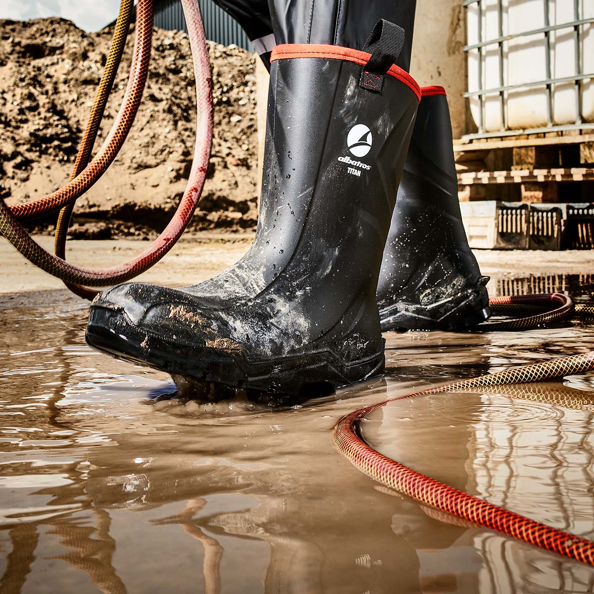 Schwarze ALBATROS® TITAN Gummistiefel mit rotem Rand, getragen in einer schlammigen Arbeitsumgebung mit Wasser und Schläuchen.
