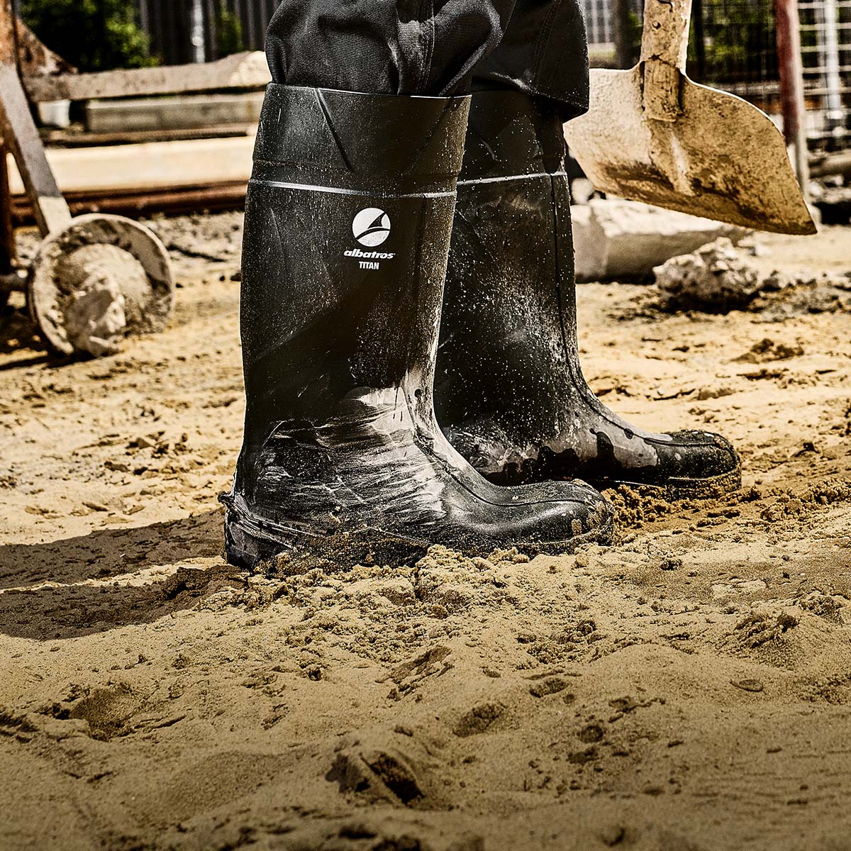 Schwarze ALBATROS® TITAN Gummistiefel mit sichtbarem Logo, getragen im sandigen Baustellenumfeld neben einer Schaufel.
