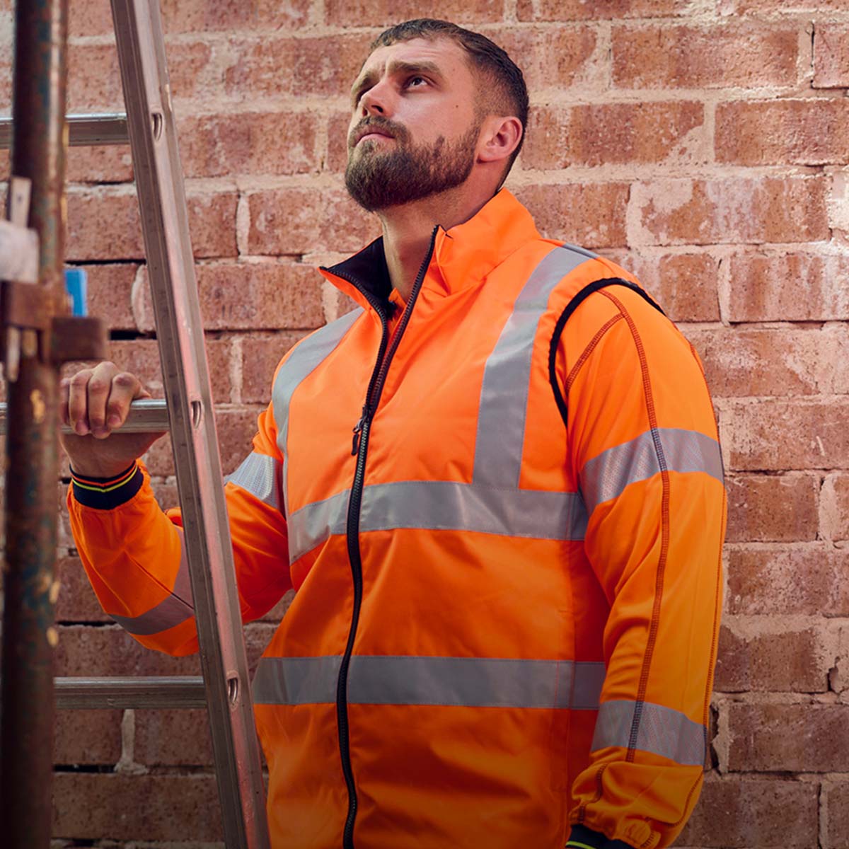Leuchtend orangefarbene Warnschutzjacke der australischen Marke BISLEY® mit silbernen Reflexstreifen, getragen von einer Person vor einer Backsteinwand neben einer Leiter.