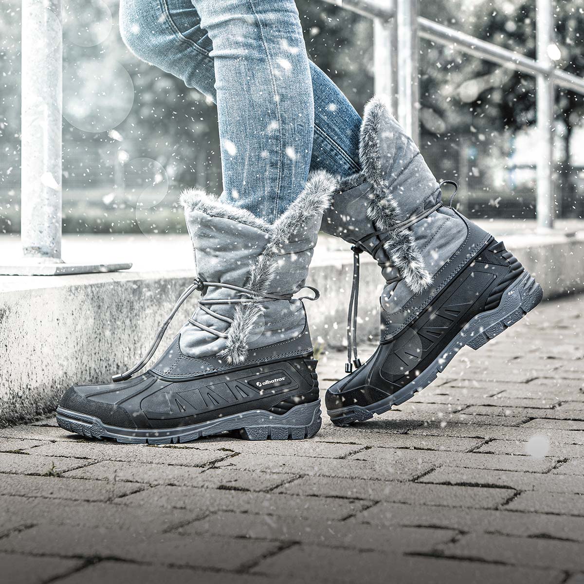 Graue ALBATROS® Winterstiefel mit Kunstfellrand und Schnürung, getragen im Schnee auf gepflastertem Untergrund.