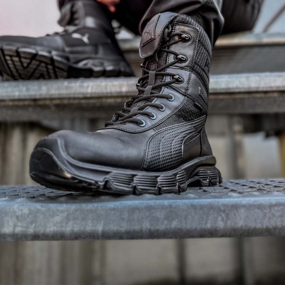 Schwarzer Einsatzstiefel von PUMA SAFETY® mit Schnürung und stark profilierter Sohle, getragen auf einer Metalltreppe.
