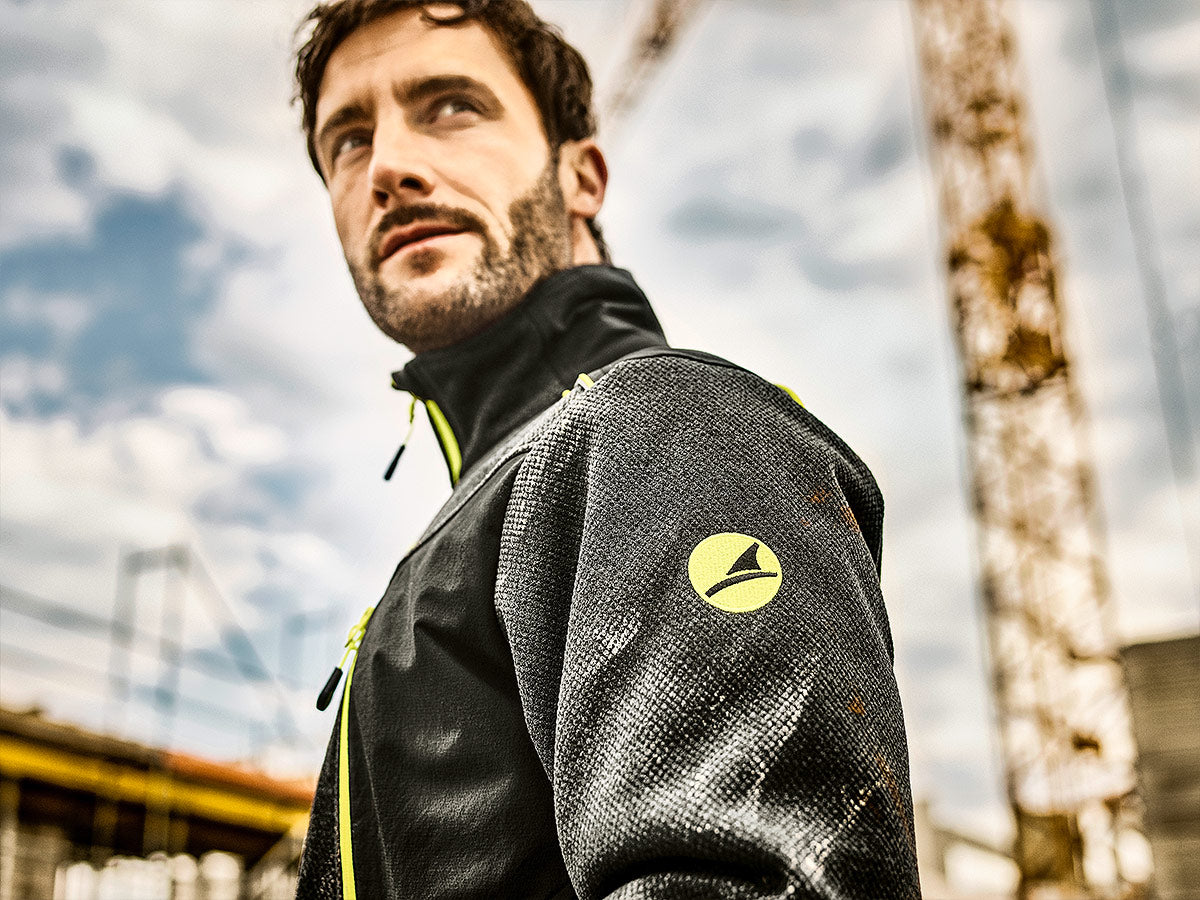 Mann auf Baustelle mit schwarzer Softshelljacke von Albatros, gelb reflektierende Details und Logo auf dem Oberarm sichtbar, im Hintergrund ein Kra und bewölkter Himmel.