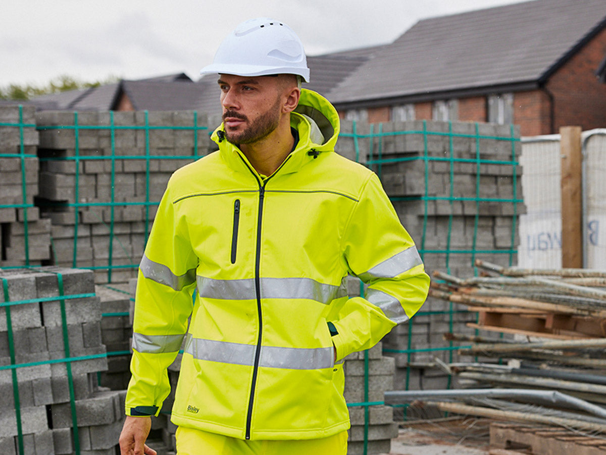 Mann mit gelber HI VIS Warnschutzjacke von Bisley und weißem Schutzhelm auf Baustelle mit Pflastersteinen und Baumaterialien im Hintergrund.