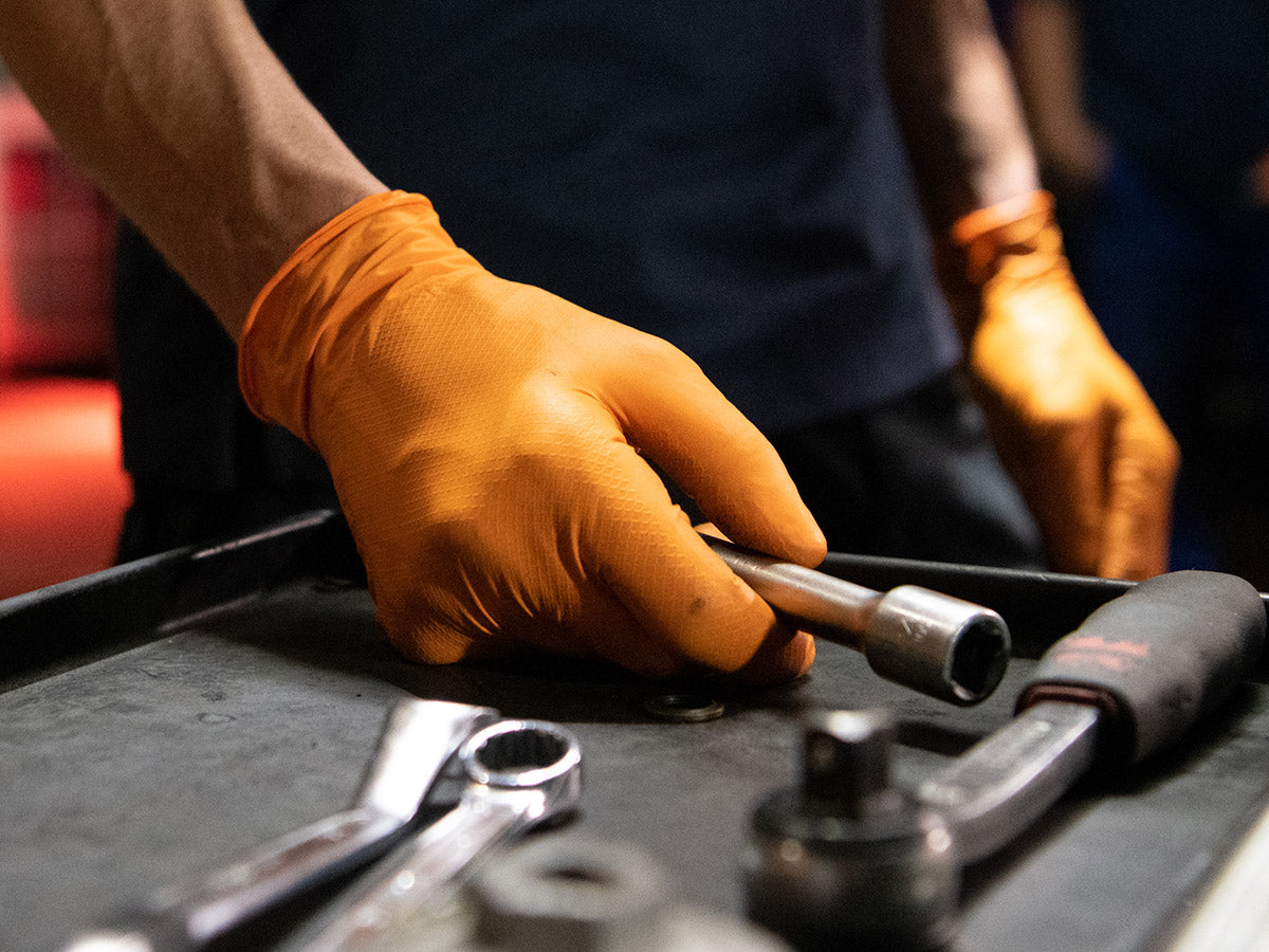 Nahaufnahme eines Mechanikers mit orangefarbenen Einweghandschuhen von Grippaz, der an einer Werkbank mit Stecknüssen und Schraubenschlüssel arbeitet.