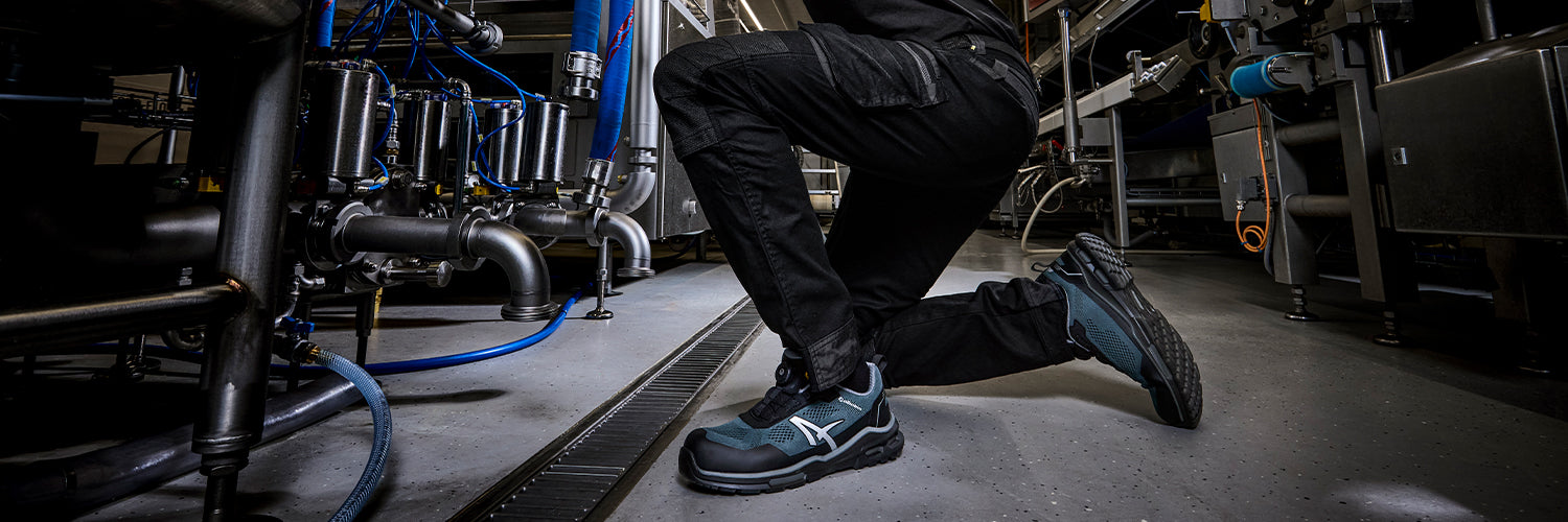 ALBATROS ECLIPSE QL LOW Sicherheitsschuh S1PS ESD in Grau und Schwarz, getragen von einer Person in kniender Position bei Wartungsarbeiten an Industrieanlagen mit Rohren, Ventilen und Kabeln auf grauem Hallenboden