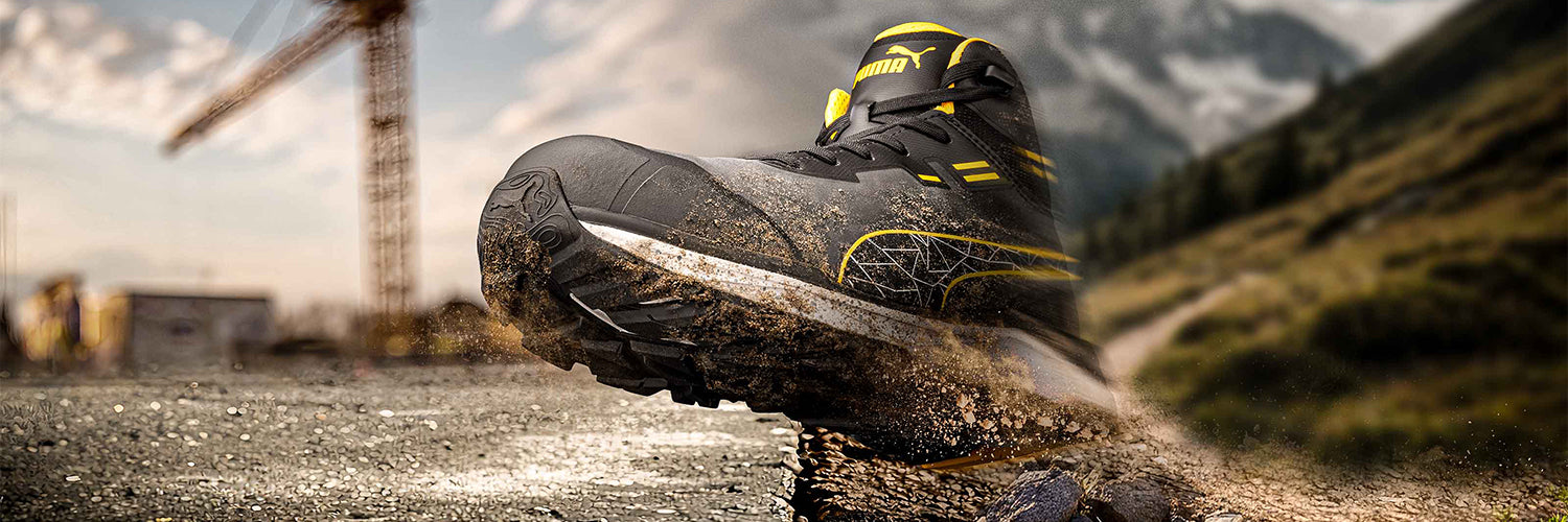 PUMA SAFETY XPLORE MID Sicherheitsschuh S3S ESD in Anthrazit mit gelben Akzenten, in Bewegung beim Auftreten auf steinigem Untergrund mit aufgewirbeltem Schmutz, vor geteilter Kulisse aus Baustelle mit Kran und Berglandschaft